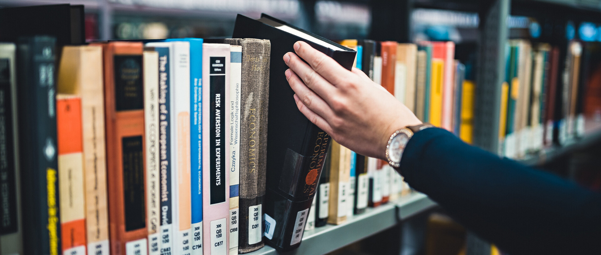 Eine Person, deren linke Hand und linker Arm im Bild sind, nimmt ein Buch aus einem Regal in der Bibliothek bzw. stellt es zurück. Die Person trägt eine Armbanduhr und einen dunkelblauen Pullover.