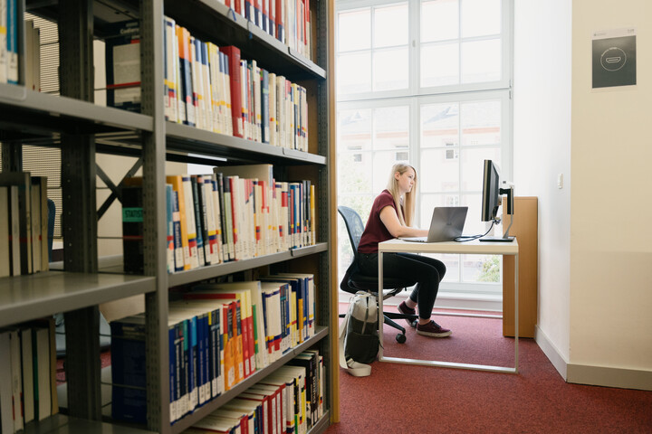 Eine junge Frau sitzt im Bibliotheksbereich Schloss Schneckenhof neben einem Bücherregal am Tisch und arbeitet am Laptop.