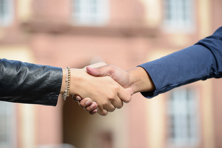 Handschlag, im Bild sind zwei Hände und jeweils ein Aussschnitt der Arme zu sehen. Die Person links im Bild trägt eine schwarze Jacke und eine goldene Armkette, die Person rechts im Bild trägt eine blaue Jacke. Im Hintergrund ist das Schloss Ehrenhof. 