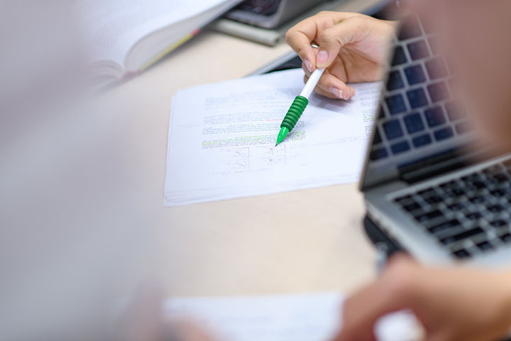 Eine Person, deren Hand im Bild ist, hält einen Stift und deutet auf ein Diagramm auf einer Buchseite. Im Hintergrund liegt ein Buch, rechts im Bild ist eine Tastatur, die sich spiegelt.